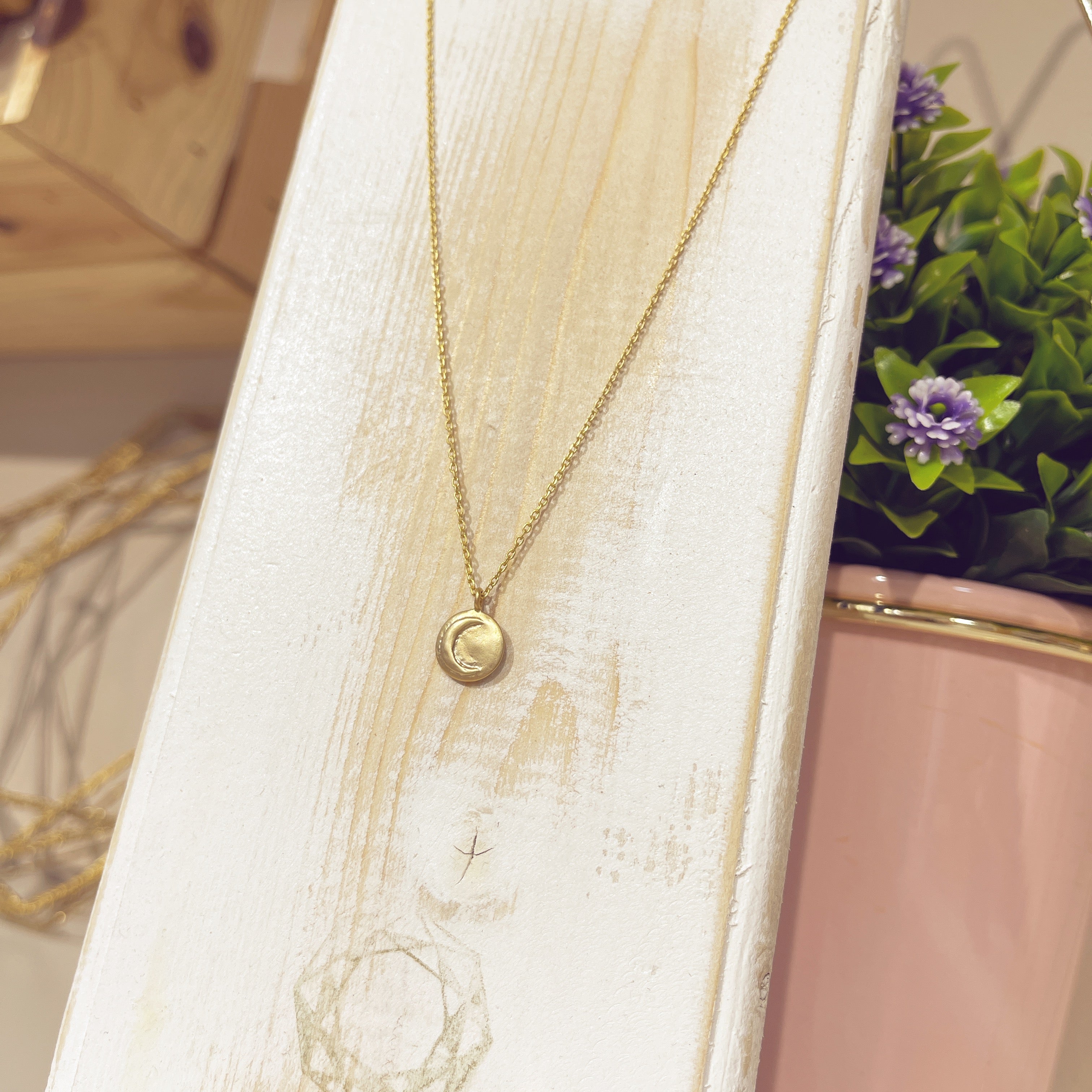 Collar medallita luna dorada sobre expositor blanco con flores de fondo