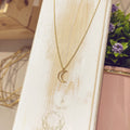 Collar luna silueta dorada sobre expositor blanco con flores de fondo