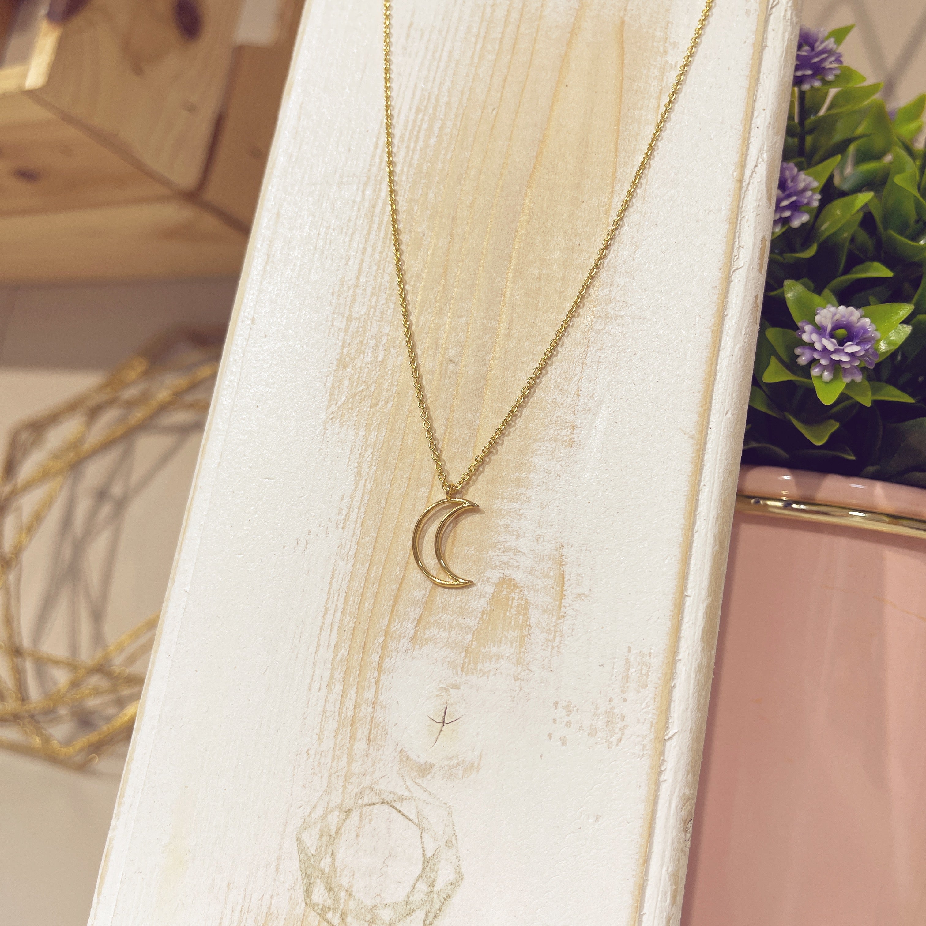 Collar luna silueta dorada sobre expositor blanco con flores de fondo