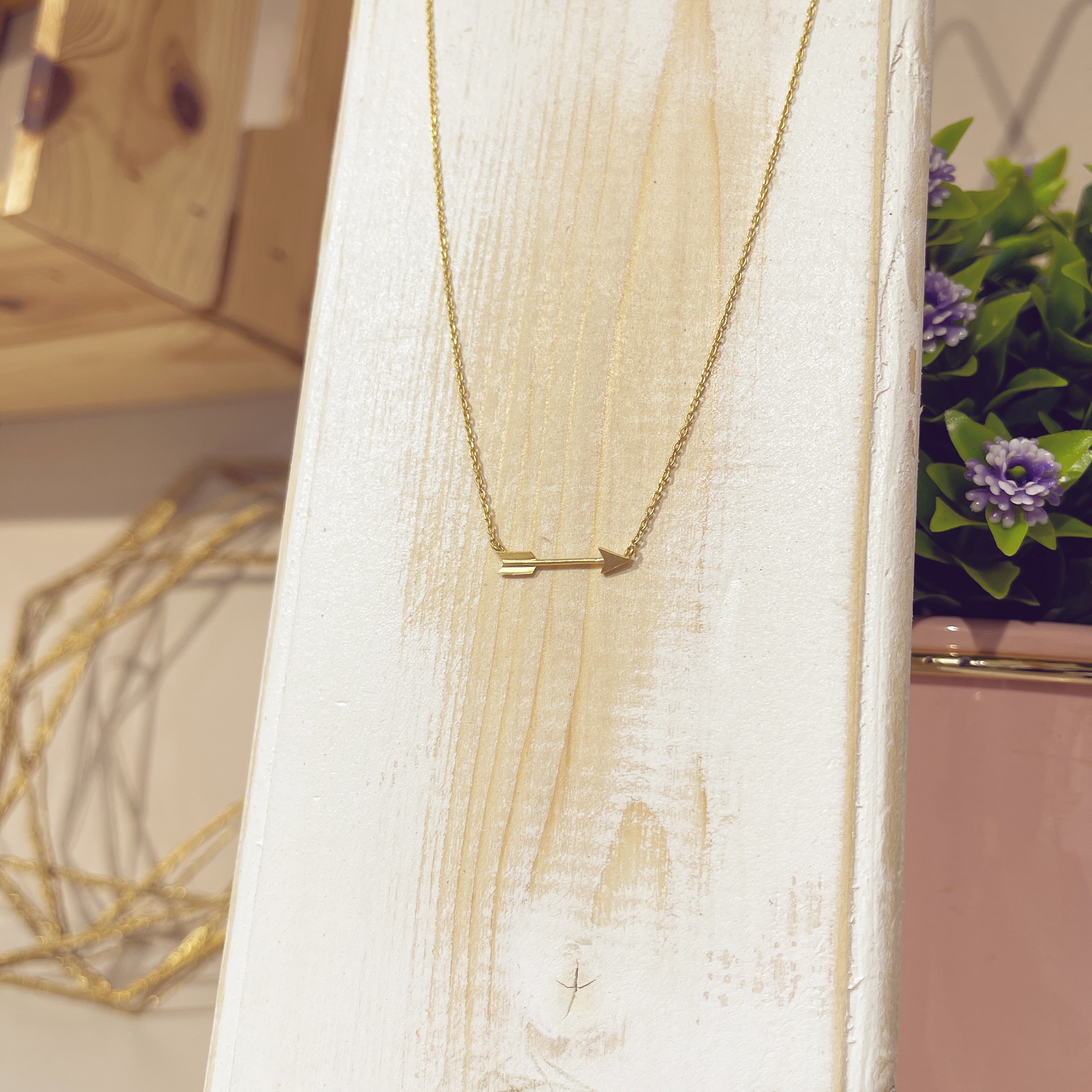 Collar flechita dorado sobre expositor blanco con flores de fondo