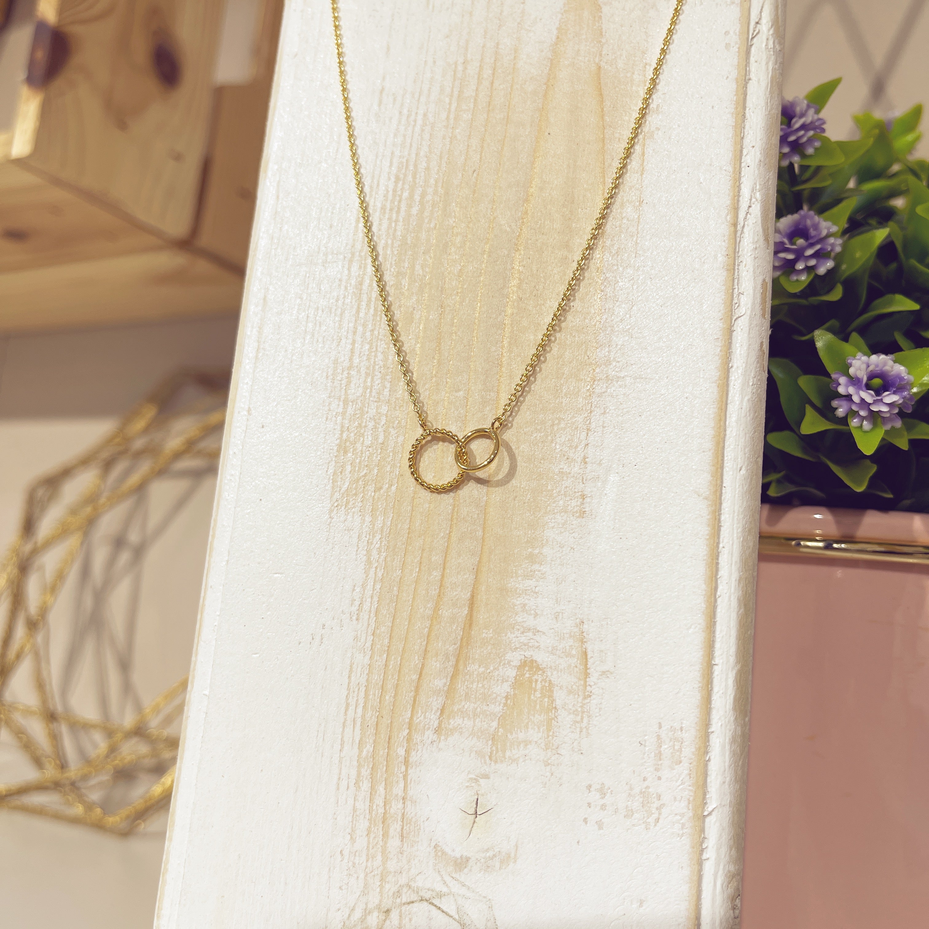 Collar arito entrelazado dorado sobre expositor blanco con flores de fondo