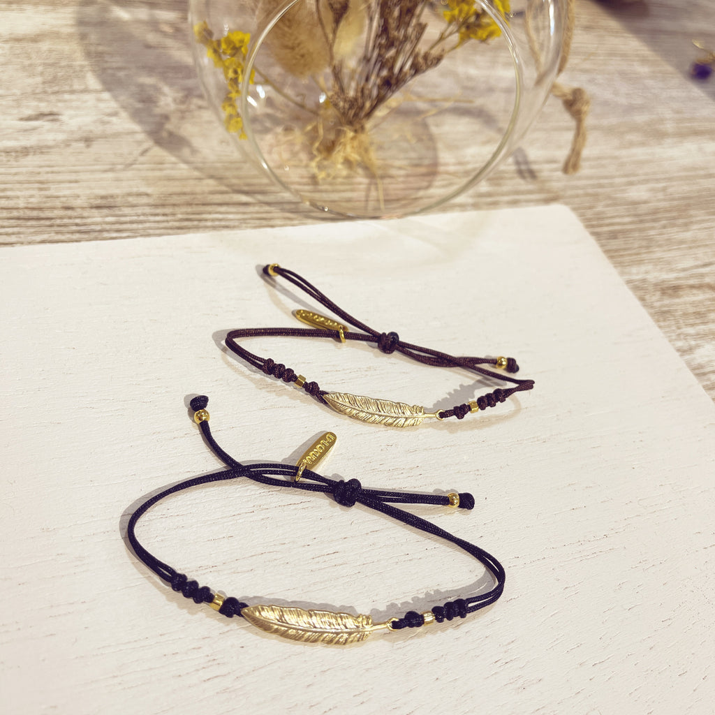 Dos pulseras Pluma Real doradas en cordón negro sobre superficie blanca