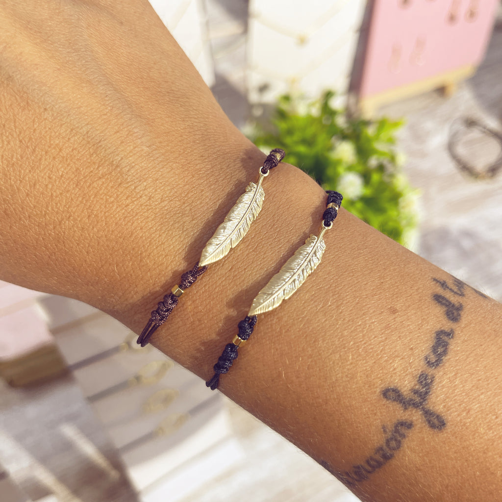 Pulseras Pluma Real doradas en cordón negro en muñeca